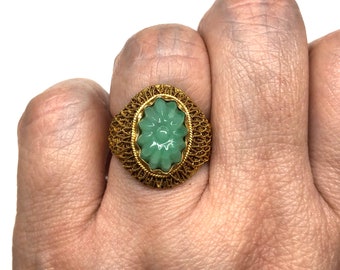 Könnte beinhalten: Ein goldfarbener Ring mit einem hellgrünen, ovalen Jade-Stein, der mit einem floralen Design geschnitzt ist. Der Jade ist in einem filigranen Rahmen gefasst. Der Ring wird an einer Hand getragen.