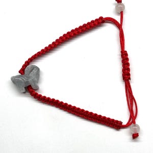 May include: A red cord bracelet featuring a gray carved stone animal charm. The bracelet is constructed with a braided knot pattern and includes an adjustable sliding closure with two small, translucent, round beads at the ends of the cords.