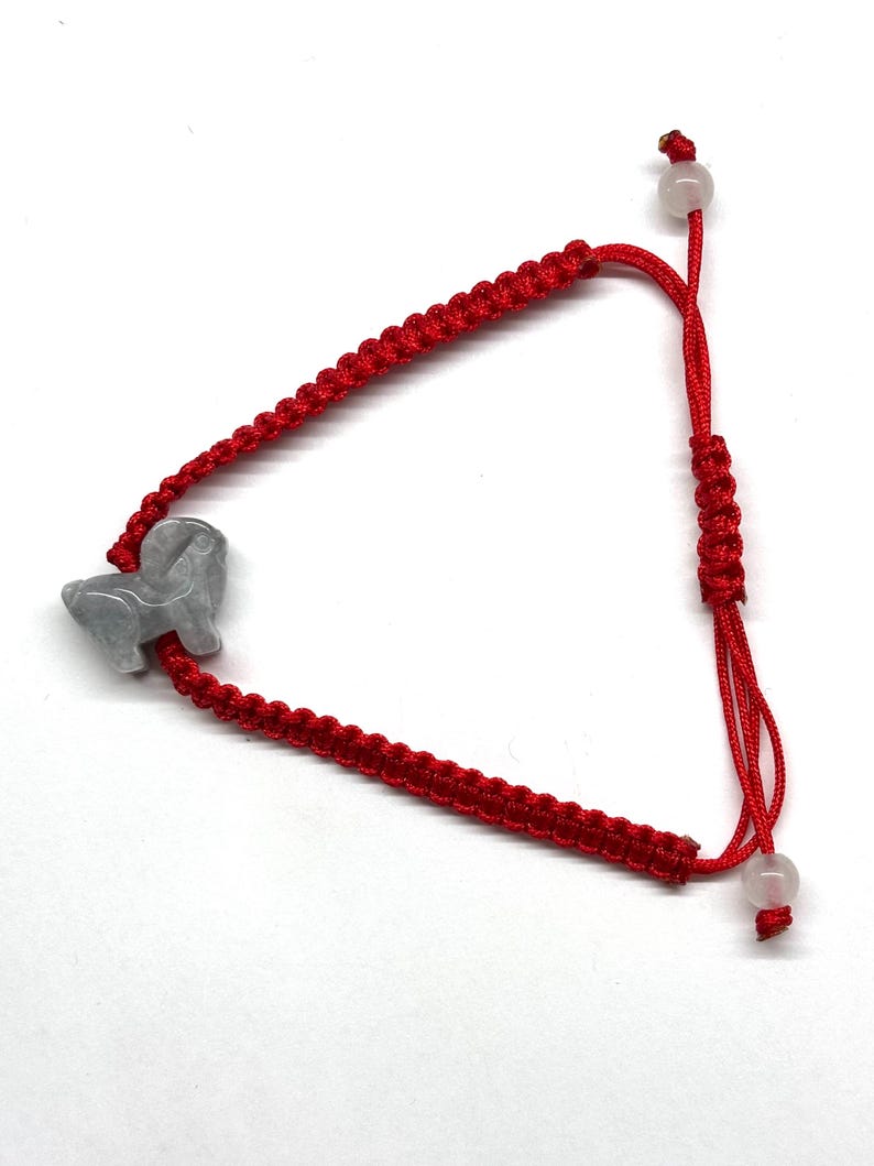 May include: A red cord bracelet featuring a gray carved stone animal charm. The bracelet is constructed with a braided knot pattern and includes an adjustable sliding closure with two small, translucent, round beads at the ends of the cords.
