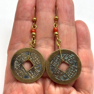 May include: A pair of antique-style earrings featuring bronze-colored coin pendants with Chinese characters. The earrings have gold-colored hooks and are accented with small red beads. The coins have a square hole in the center.