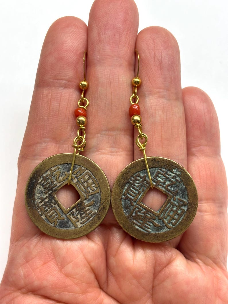 May include: A pair of antique-style earrings featuring bronze-colored coin pendants with Chinese characters. The earrings have gold-colored hooks and are accented with small red beads. The coins have a square hole in the center.