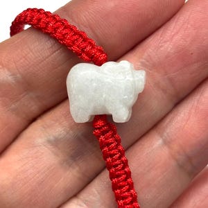 May include: A bracelet featuring a white, carved stone pig charm centred on a vibrant red braided cord. The pig figure is smooth and opaque, contrasting with the textured, woven red string that forms the adjustable band of this wrist accessory.