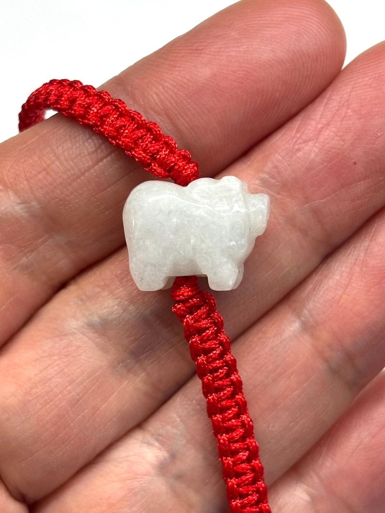May include: A bracelet featuring a white, carved stone pig charm centred on a vibrant red braided cord. The pig figure is smooth and opaque, contrasting with the textured, woven red string that forms the adjustable band of this wrist accessory.