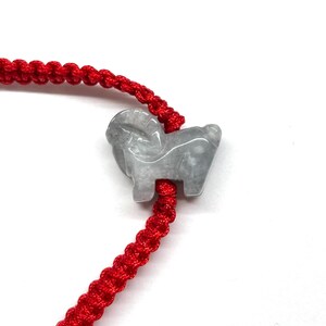 May include: A bracelet featuring a bright red braided cord and a carved light gray stone charm in the shape of a ram. The ram charm is positioned along the center of the cord, which displays a consistent woven pattern throughout its length.