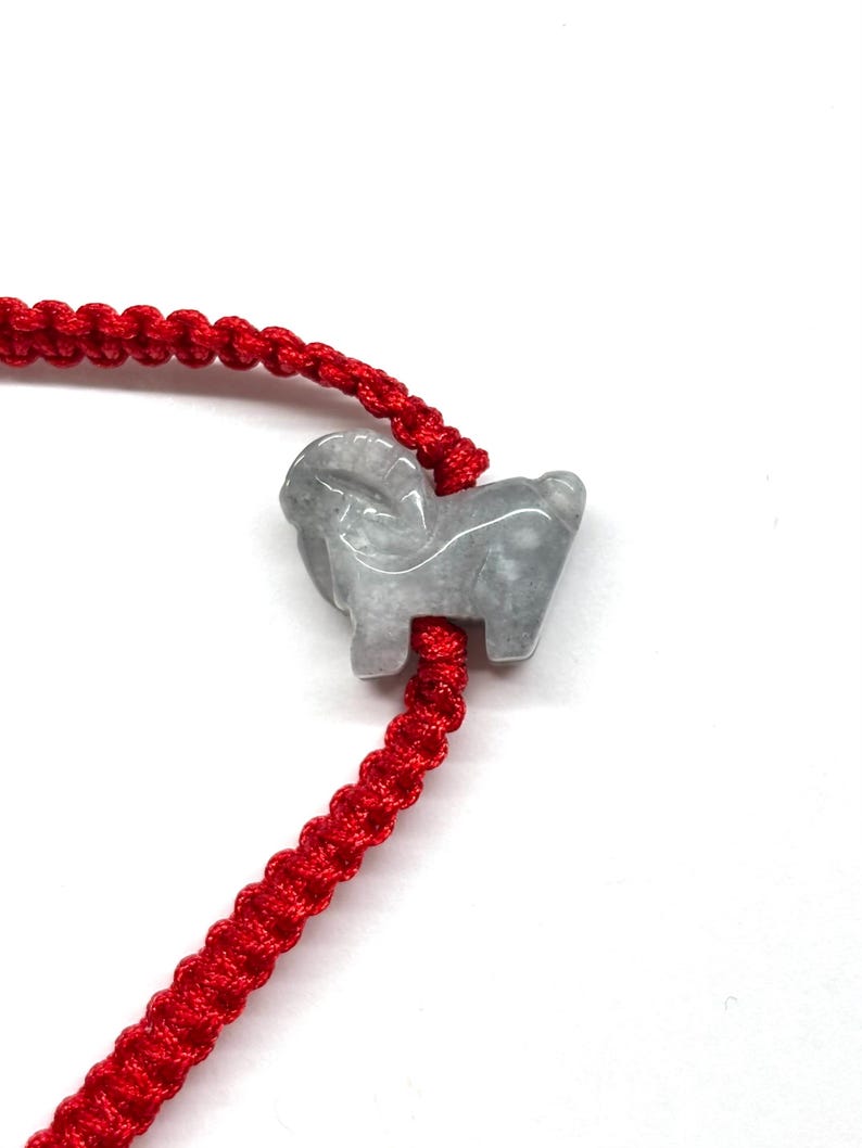 May include: A bracelet featuring a bright red braided cord and a carved light gray stone charm in the shape of a ram. The ram charm is positioned along the center of the cord, which displays a consistent woven pattern throughout its length.