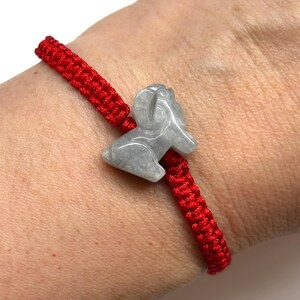 May include: A bright red woven cord bracelet featuring a central light gray stone charm carved into the shape of a ram. The bracelet is adjustable and sits securely on the wrist, showcasing a smooth, polished finish on the stone animal figure.