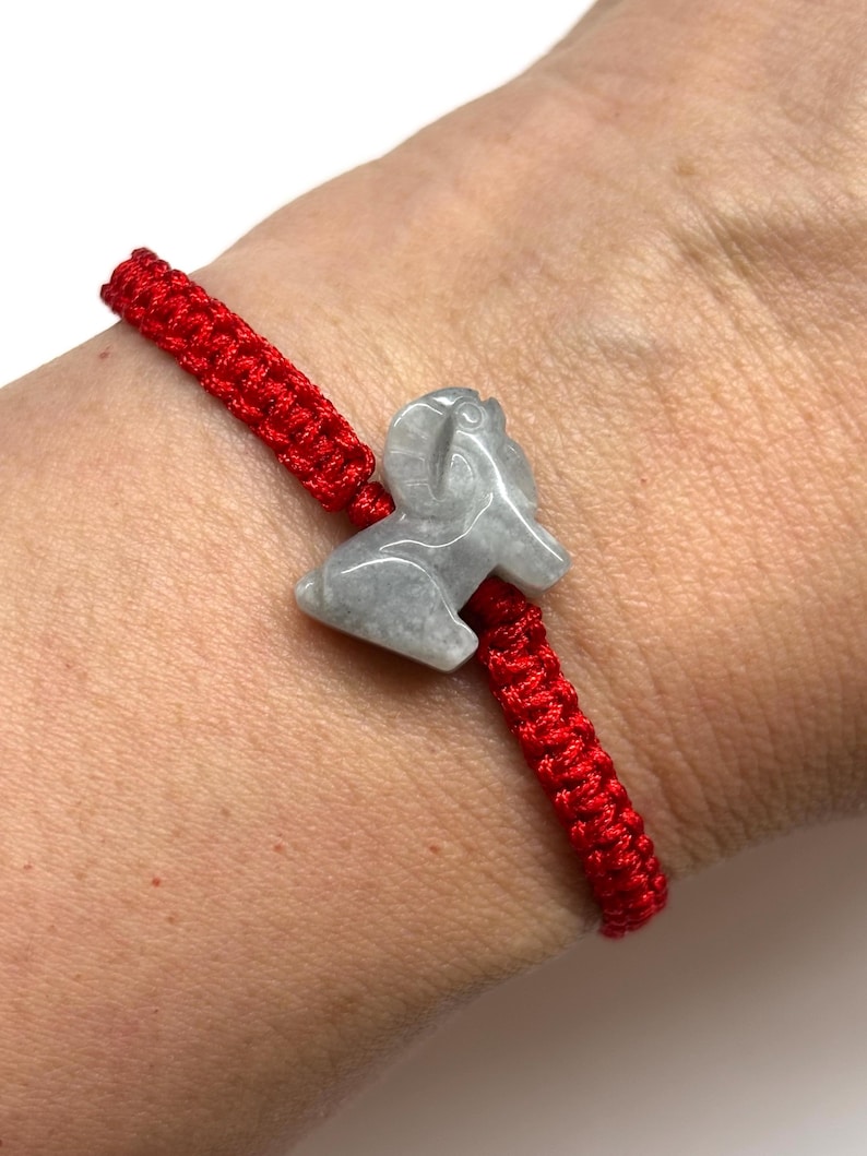 May include: A bright red woven cord bracelet featuring a central light gray stone charm carved into the shape of a ram. The bracelet is adjustable and sits securely on the wrist, showcasing a smooth, polished finish on the stone animal figure.