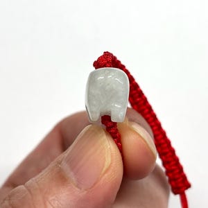 May include: A small, translucent, pale grey stone bead carved into the shape of a stylised animal, threaded onto a vibrant red braided cord bracelet. The bead features a smooth, polished surface with subtle internal veining and a rounded top.