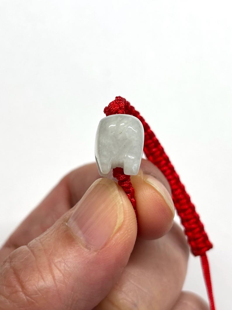May include: A small, translucent, pale grey stone bead carved into the shape of a stylised animal, threaded onto a vibrant red braided cord bracelet. The bead features a smooth, polished surface with subtle internal veining and a rounded top.