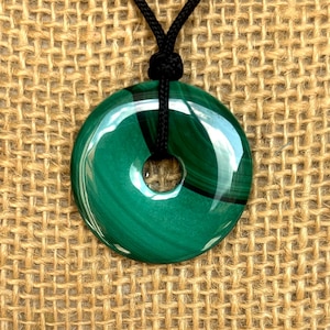 May include: A green malachite stone pendant in the shape of a circle with a hole in the center. The pendant is on a black cord.