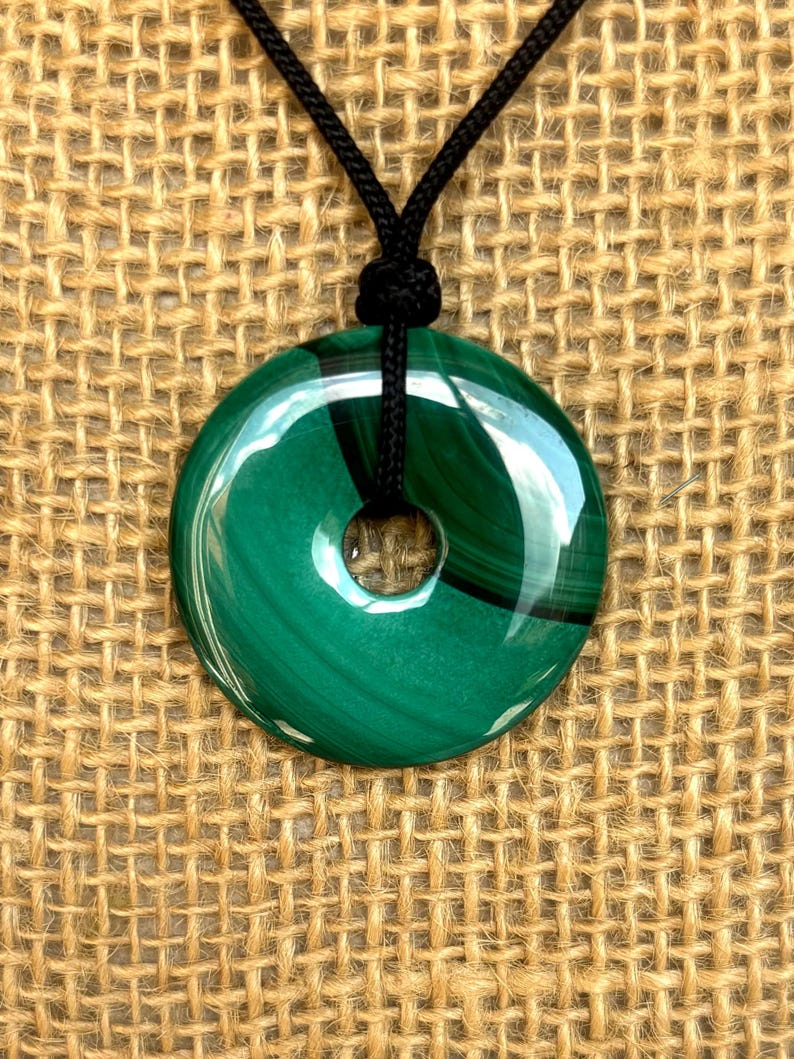 May include: A green malachite stone pendant in the shape of a circle with a hole in the center. The pendant is on a black cord.