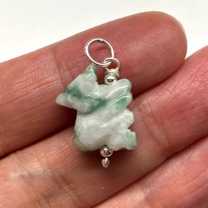 May include: A jade pendant carved in the shape of a mythical creature, with white and green hues. The pendant is attached to a silver-colored metal loop and a small bead. The pendant is held in a person's hand.