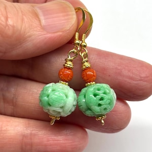 May include: A pair of gold-toned earrings featuring carved green jade spheres with floral designs. The earrings also have small orange beads and gold-colored hooks. The earrings are held up by a person's hand.