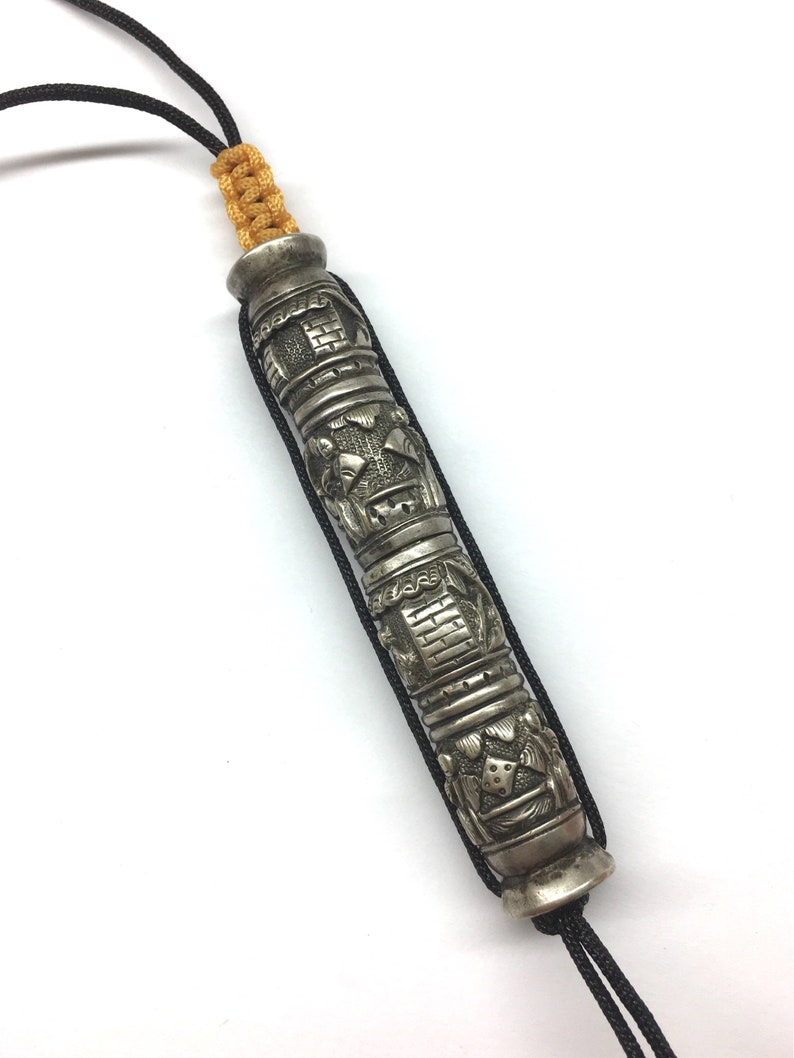 May include: A silver prayer wheel with intricate carvings and a black cord. The prayer wheel is decorated with a repeating pattern of symbols and figures.