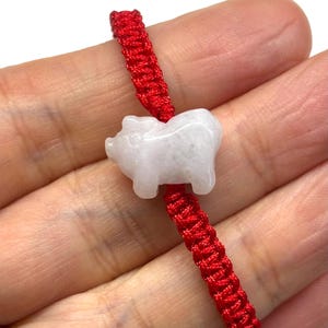 May include: A bracelet featuring a carved, pale white, translucent stone pig charm centred on a vibrant red braided cord. The pig figure is smooth and rounded, showcasing a simple silhouette. The red cord is woven in a repeating knot pattern.