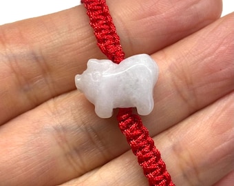 May include: A bracelet featuring a carved, pale white, translucent stone pig charm centered on a vibrant red braided cord. The pig figure is smooth and rounded, showcasing a simple silhouette. The red cord is woven in a repeating knot pattern.