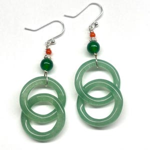 May include: A pair of green jade earrings with silver hooks. Each earring features two interlocking circles with a green jade bead and an orange bead hanging from the top.