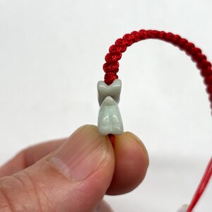 May include: A bracelet featuring a bright red braided cord and a small, pale green carved stone bead shaped like a stylized animal figure. The smooth, light green stone bead is threaded onto the vibrant red cord, which has a knotted texture.