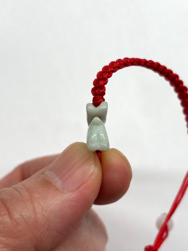 May include: A bracelet featuring a bright red braided cord and a small, pale green carved stone bead shaped like a stylized animal figure. The smooth, light green stone bead is threaded onto the vibrant red cord, which has a knotted texture.