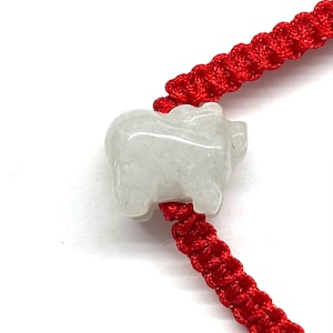 May include: A bracelet featuring a bright red braided cord and a single, carved, translucent white stone charm in the shape of a pig. The charm is positioned centrally on the cord, which displays a consistent, repeating knot pattern throughout.
