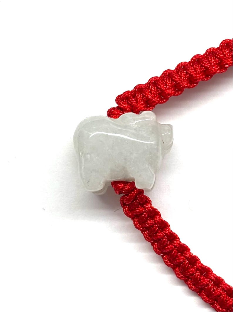 May include: A bracelet featuring a bright red braided cord and a single, carved, translucent white stone charm in the shape of a pig. The charm is positioned centrally on the cord, which displays a consistent, repeating knot pattern throughout.