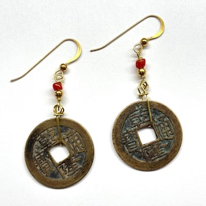 May include: A pair of gold-colored earrings featuring bronze-colored coin pendants with square cutouts and Chinese characters. Each earring has a small strand of red beads and gold-colored ear hooks.