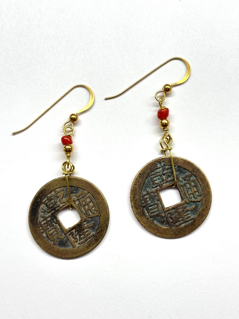 May include: A pair of gold-colored earrings featuring bronze-colored coin pendants with square cutouts and Chinese characters. Each earring has a small strand of red beads and gold-colored ear hooks.