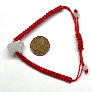 May include: A red braided cord bracelet featuring a light grey carved stone animal charm and two small round beads at the adjustable closure. A Canadian one-cent coin is placed next to the bracelet for scale, showing the profile of Queen Elizabeth II.