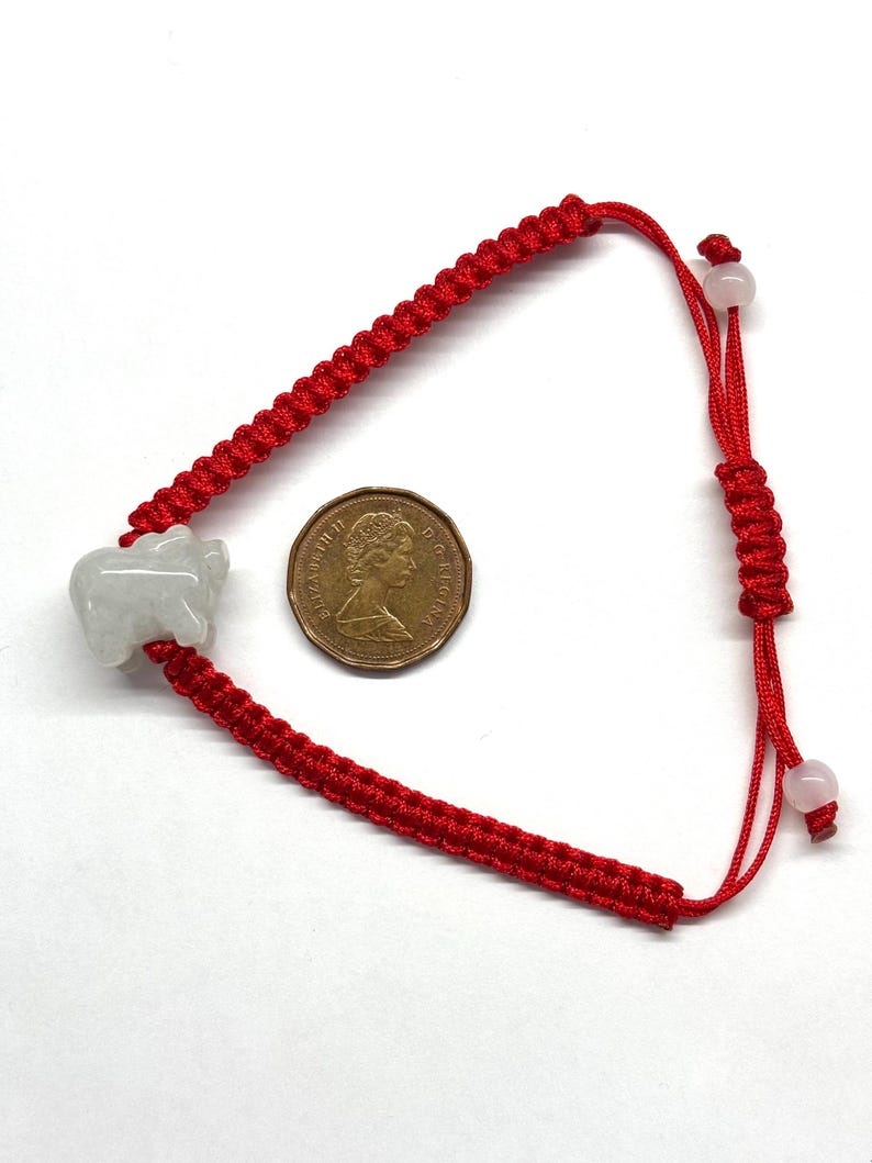May include: A red braided cord bracelet featuring a light grey carved stone animal charm and two small round beads at the adjustable closure. A Canadian one-cent coin is placed next to the bracelet for scale, showing the profile of Queen Elizabeth II.