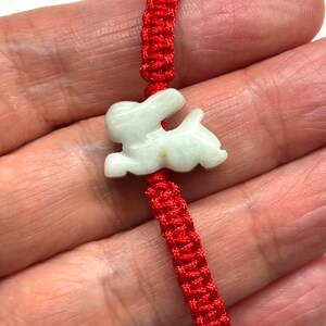 May include: A bracelet featuring a pale green, carved stone rabbit charm centered on a vibrant red woven cord. The rabbit is depicted in a leaping or running pose with smooth, polished surfaces. The cord is constructed with a braided knot pattern.