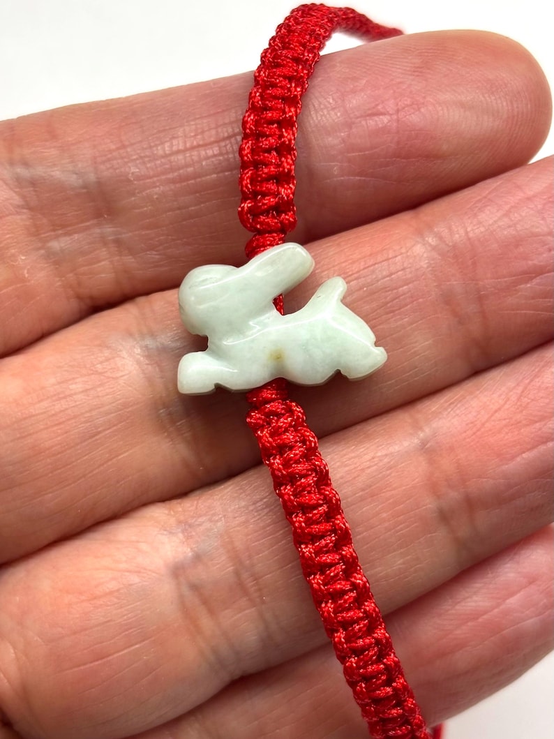 May include: A bracelet featuring a pale green, carved stone rabbit charm centered on a vibrant red woven cord. The rabbit is depicted in a leaping or running pose with smooth, polished surfaces. The cord is constructed with a braided knot pattern.