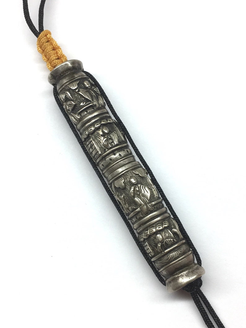 May include: A silver prayer wheel with intricate carvings and a black cord. The wheel is decorated with raised figures and designs.
