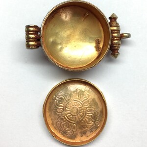 K&ouml;nnte beinhalten: Ein goldfarbener Metallbeh&auml;lter mit einem Scharnierdeckel. Der Deckel hat ein dekoratives Design und der Beh&auml;lter ist leer.