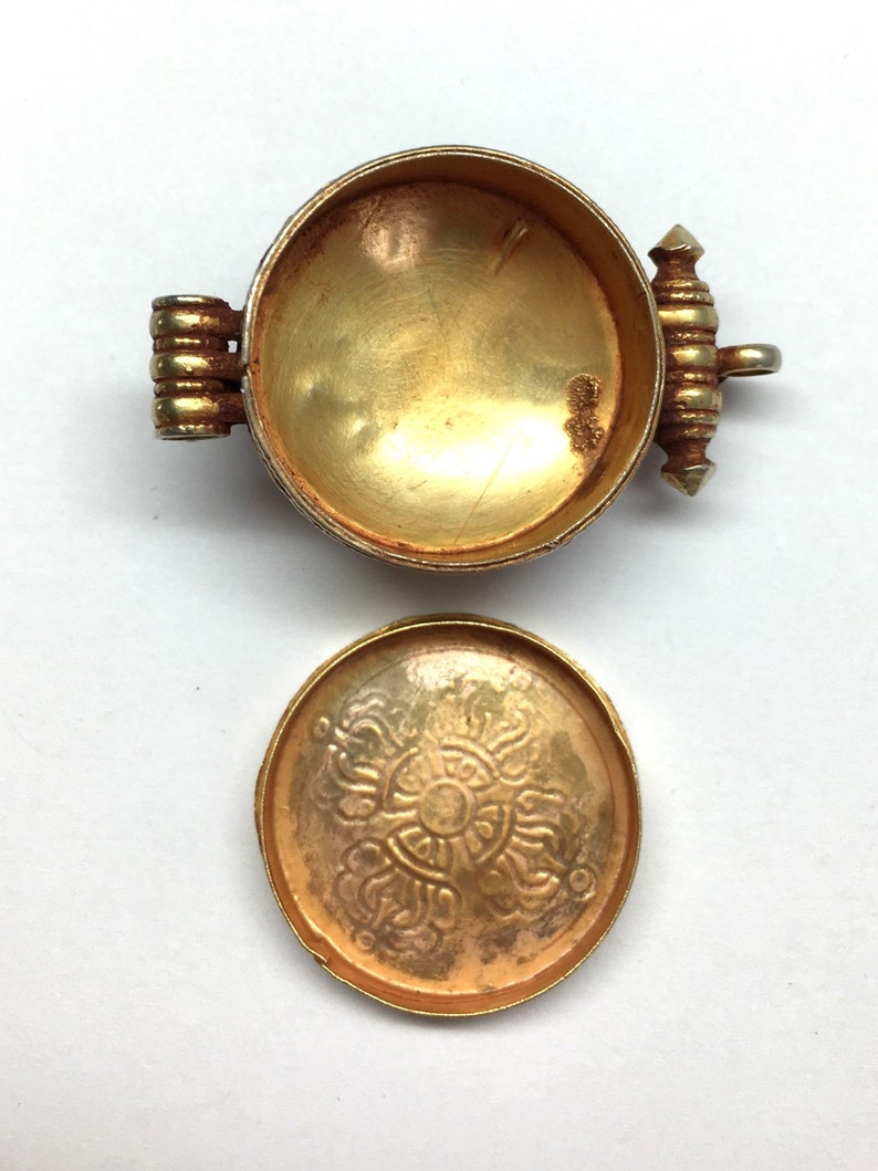 K&ouml;nnte beinhalten: Ein goldfarbener Metallbeh&auml;lter mit einem Scharnierdeckel. Der Deckel hat ein dekoratives Design und der Beh&auml;lter ist leer.