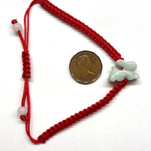 May include: A red braided cord bracelet featuring a pale green carved jade rabbit charm. The bracelet includes an adjustable sliding knot closure with two small light-colored beads at the ends, positioned next to a Canadian one-cent coin for scale.