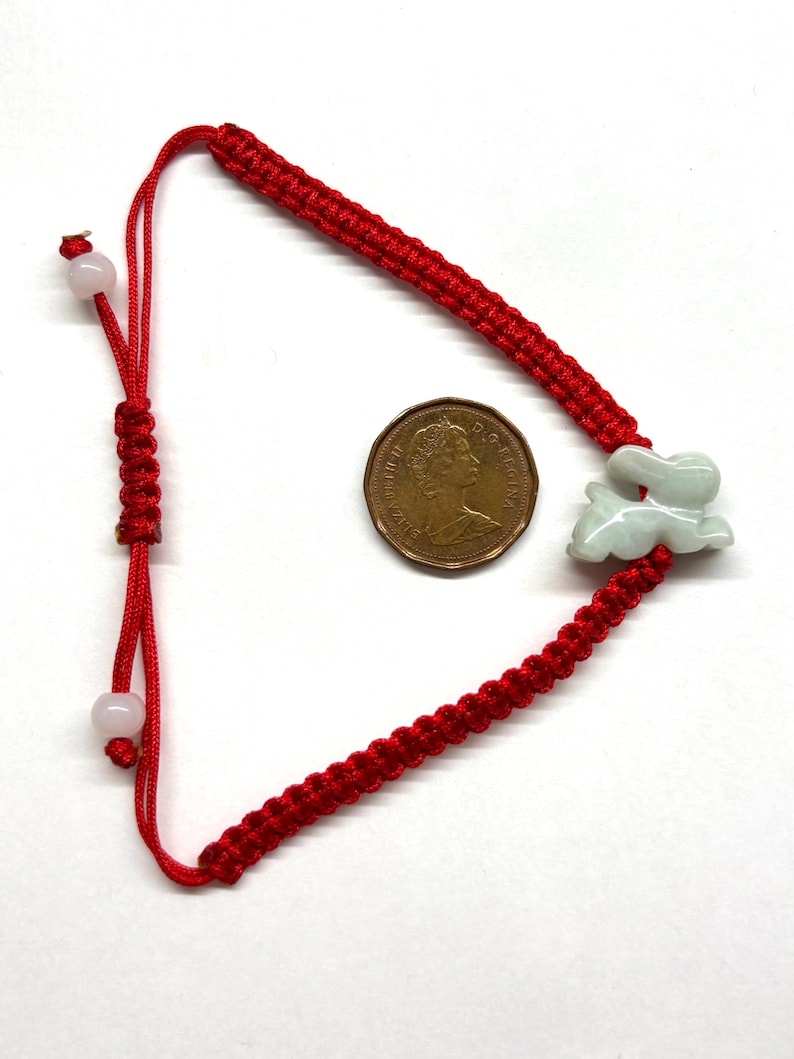 May include: A red braided cord bracelet featuring a pale green carved jade rabbit charm. The bracelet includes an adjustable sliding knot closure with two small light-colored beads at the ends, positioned next to a Canadian one-cent coin for scale.