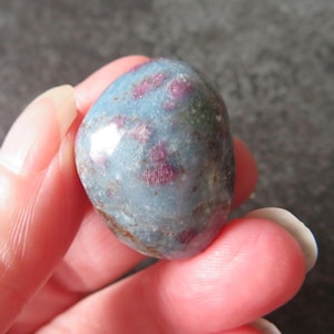 May include: A polished gemstone with a rounded shape. The stone has a light blue base color with patches of deep red and brown. The gemstone is held between fingers.