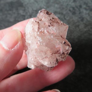 May include: A small, translucent quartz crystal cluster featuring pale pink hues and distinct dark reddish-brown mineral inclusions scattered across its faceted, angular surface. The mineral specimen displays a jagged, multi-pointed structure.