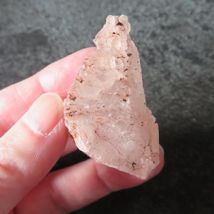 May include: A translucent, pale pink crystal fragment with an irregular, jagged shape and rough, textured surfaces. The mineral specimen features small, dark brown inclusions scattered across its crystalline structure and uneven edges.