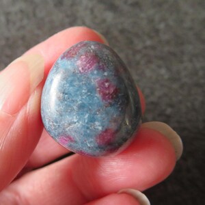 May include: A smooth, oval gemstone with a mottled appearance. The stone displays a mix of blue-grey and deep red-purple colours, creating a unique, speckled pattern. The polished surface reflects light, highlighting the colour variations.