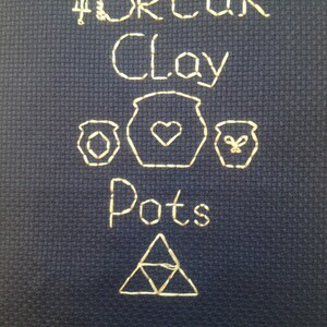 May include: A cross-stitch design on a dark blue fabric with the words "Break Clay Pots" and a stylized Triforce symbol.