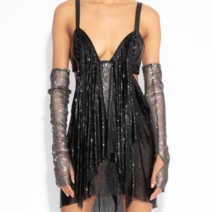 Puede incluir: Un vestido negro de lentejuelas corto con escote pronunciado y falda de tul con volantes. El vestido tiene mangas largas transparentes con pedrería.