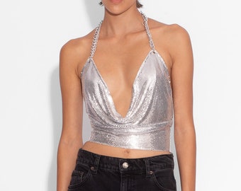 May include: A silver chainmail halter top with a deep V-neckline. The top is cropped and has a fitted silhouette. The model is wearing the top with black jeans.