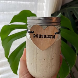 May include: A clear glass jar filled with a creamy, off-white substance, held in a person's hand. The jar has a silver lid and a wooden heart-shaped tag that reads "Doughlene" in black letters. The background includes green leaves and a white curtain.