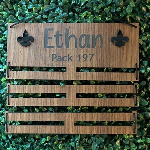 May include: A rectangular wooden display board with a dark wood grain finish, featuring the engraved text "Ethan" and "Pack 197". The board includes two black fleur-de-lis accents and three horizontal slots connected by black cords for hanging items.