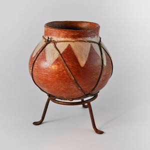 Peut inclure: Un pot en terre cuite avec des motifs géométriques blancs et une bande tissée brune autour du bord. Le pot est posé sur un support en métal à trois pieds.