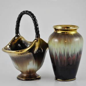 May include: A pair of ceramic vases with a brown, gold and green glaze. The vase on the left has a black rope handle and is shaped like a basket. The vase on the right is a traditional cylinder shape.
