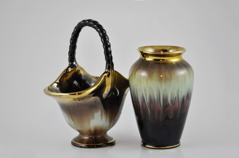 May include: A pair of ceramic vases with a brown, gold and green glaze. The vase on the left has a black rope handle and is shaped like a basket. The vase on the right is a traditional cylinder shape.