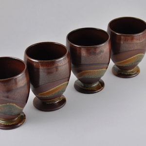May include: Four brown ceramic cups with a brown, green and yellow glaze pattern. The cups have a rounded base and a wide opening.
