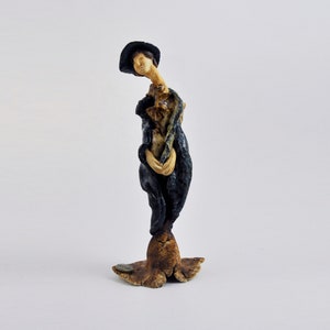 Peut inclure: Une figurine en céramique d'une personne portant un chapeau noir et une robe bleue. La figurine est debout sur une base brune.